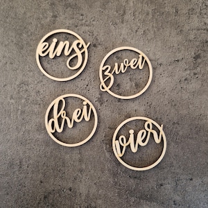 Puede incluir: Cuatro círculos de madera con las palabras alemanas "eins", "zwei", "drei" y "vier" escritas en una fuente cursiva.