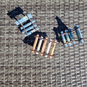 May include: Three small wooden ladder-like structures, each holding several rolled-up paper banknotes. The structures are arranged in a triangular formation, with one featuring blue-toned notes and two featuring orange-toned notes.