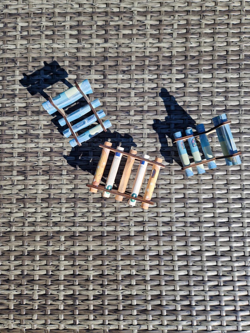 May include: Three small wooden ladder-like structures, each holding several rolled-up paper banknotes. The structures are arranged in a triangular formation, with one featuring blue-toned notes and two featuring orange-toned notes.