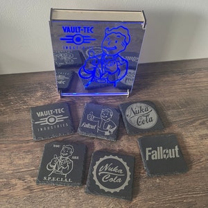 Könnte beinhalten: Sechs schwarze Schieferuntersetzer mit verschiedenen Fallout- und Nuka-Cola-Designs. Die Untersetzer zeigen das Vault-Tec-Logo, das Fallout-Logo und das Nuka-Cola-Logo. Ein Untersetzer zeigt den Text "You are S.P.E.C.I.A.L."