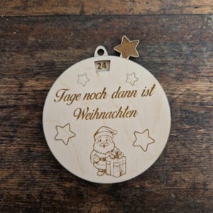 May include: A wooden Christmas ornament with the text "Tage noch dann ist Weihnachten" and a Santa Claus illustration. The ornament has a star-shaped cutout and a small window displaying the number 24. The ornament is round with star-shaped outlines.