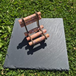 May include: A miniature chair constructed from dark brown wooden side supports and horizontal slats made of rolled orange paper currency featuring white star patterns. The small furniture piece is positioned on a square, dark gray slate tile.
