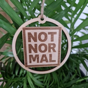 Könnte beinhalten: Ein runder Holzanhänger mit einem quadratischen Ausschnitt in der Mitte. Der Ausschnitt zeigt den Text "NOT NORMAL" in dunkelbrauner Farbe.