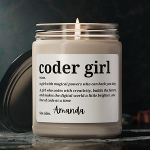 May include: A beige candle with a glass jar and a black lid. The label reads "coder girl" and defines it as "a girl with magical powers who can hack you day. A girl who codes with creativity, builds the future, and makes the digital world a little brighter, one line of code at a time. See also. Amanda"