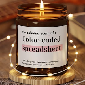 May include: A lit candle in a brown glass jar with a label that reads "the calming scent of a Color-coded spreadsheet." The candle sits on a small wooden coaster, surrounded by string lights. A guitar and book are in the background.