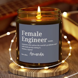 May include: A lit candle in a glass jar with a label that reads "Female Engineer." The label is dark gray with white text and the name "Amanda" in cursive. The candle sits on a wooden coaster, with a guitar and a book in the background.