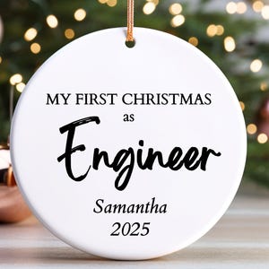 May include: A white, circular Christmas decoration with the words "MY FIRST CHRISTMAS as Engineer" in black. The name "Samantha" and the year "2025" are also printed on the ornament. A gold string is attached for hanging.