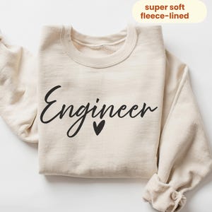 May include: A cream-coloured fleece-lined sweatshirt with the word "Engineer" in black script, a small black heart below the word, and the text "super soft fleece-lined" in a yellow oval.