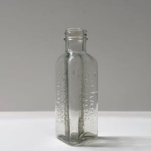 May include: A clear, antique glass bottle with embossed text. The bottle has a rectangular shape with rounded shoulders and a narrow neck. The text is partially visible and appears to be a label. The bottle is empty and sits on a white surface.