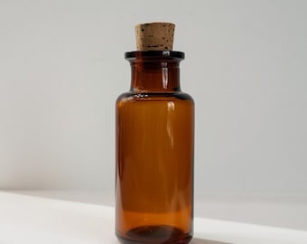 May include: A cylindrical amber glass bottle with a smooth, translucent finish and a wide neck. The bottle is sealed with a light brown, porous cork stopper that fits snugly into the opening, creating a simple and functional storage container.
