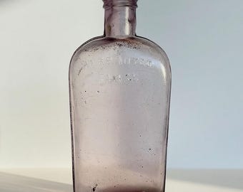 May include: A vintage light purple glass flask with a rounded shoulder and a short neck. The glass features embossed text that reads WARRANTED FLASK across the upper body. The surface shows small air bubbles and subtle imperfections in the glass.