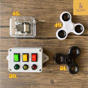 May include: A clear, rectangular music box with a silver crank and internal mechanism. A white and black fidget spinner and a black fidget spinner are also shown. A white box with green, yellow, and red switches and lights is also shown.