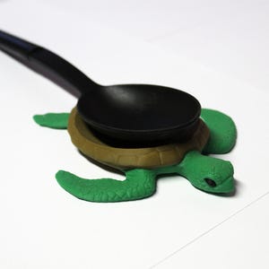 Può includere: Un cucchiaio nero poggia su un poggiacucchiaio a forma di tartaruga verde e marrone. Il poggiacucchiaio presenta un design a guscio di tartaruga ed è realizzato in un materiale resistente. Il cucchiaio è in plastica nera.