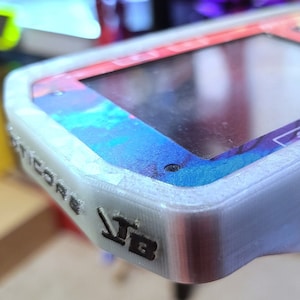 Op de afbeelding: Een witte 3D-geprinte behuizing voor een klein elektronisch apparaat. De behuizing heeft een textuur en de woorden "V1B" en "MICRO" zijn aan de zijkant geprint. Het apparaat heeft een kleurrijk scherm en een paar knoppen aan de zijkant.