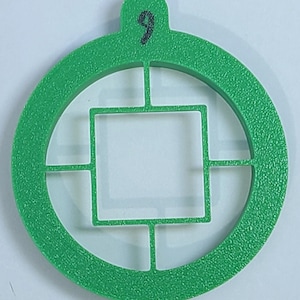 Pode incluir: Círculo de plástico verde com um quadrado cortado no centro. O círculo tem um pequeno laço na parte superior com o número "9" escrito nele.