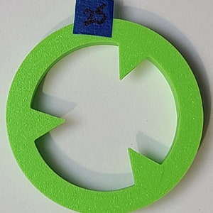 Pode incluir: Um anel de plástico verde brilhante com quatro recortes em forma de flecha. O anel tem um adesivo azul com o número "25" escrito em preto.