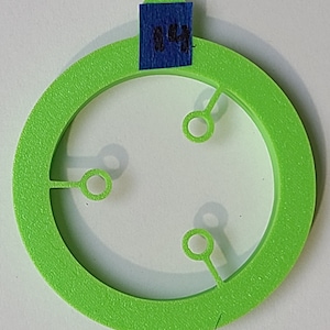 Może przedstawiać: Jasnozielony plastikowy okrąg z trzema małymi otworami i większym okręgiem wyciętym w środku. Okrąg ma szorstką fakturę.
