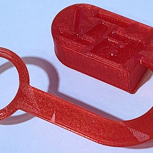 May include: A red 3D printed plastic hook with a ring on one end and a rectangular block on the other end. The block has a textured surface with a pattern.