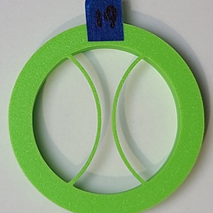 May include: A green plastic cookie cutter shaped like a tennis ball. The cutter has a raised rim and a cut-out design of a tennis ball.