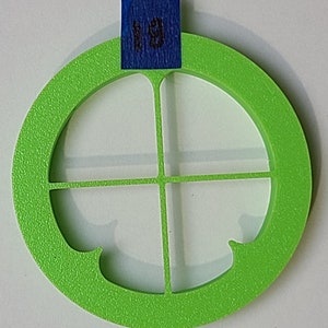 Pode incluir: Um círculo de plástico verde brilhante com um recorte em forma de cruz no centro. O círculo tem uma superfície texturizada e uma borda ligeiramente elevada.