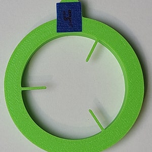 Pode incluir: Um anel de plástico verde brilhante com quatro recortes igualmente espaçados. O anel tem um adesivo azul com o número "4" escrito nele.