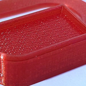 May include: A red plastic 3D printed object with a rectangular shape and a rounded top. The object has a textured surface.