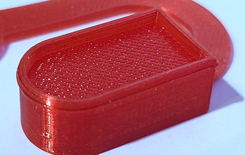 May include: A red plastic 3D printed object with a rectangular shape and a rounded top. The object has a textured surface.