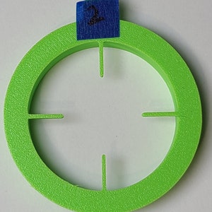 Pode incluir: Um círculo de plástico verde brilhante com quatro recortes, que se assemelha a um alvo. O círculo tem uma superfície texturizada e um pequeno rótulo azul com o número "2" escrito nele.