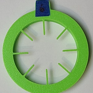 Pode incluir: Um anel de plástico verde com oito fendas igualmente espaçadas ao redor do centro. O anel tem uma superfície texturizada.