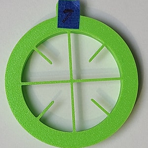 Pode incluir: Um círculo de plástico verde brilhante com um design recortado no centro. O design recortado é um círculo com quatro raios que irradiam do centro. O círculo tem uma superfície texturizada.