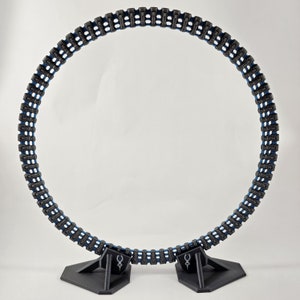 Op de afbeelding: Een zwart-blauwe 3D-geprinte ring met een textuur. De ring staat op een zwarte standaard met twee poten.