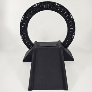 May include: A black plastic model of a Stargate, a circular portal device from the science fiction television series Stargate SG-1. The model is mounted on a black plastic base.