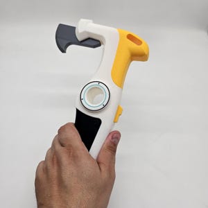 Könnte beinhalten: Ein mehrfarbiges Werkzeug mit weißem, gelbem und schwarzem Design. Das Werkzeug hat eine gebogene Klinge und einen Griff mit kreisförmigem Design. Das Werkzeug wird in einer Hand gehalten.