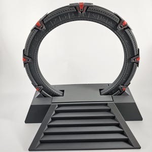 Op de afbeelding: Een 3D-geprint model van een Stargate, een fictief apparaat dat wordt gebruikt voor interstellaire reizen. De Stargate is zwart met rode accenten en is gemonteerd op een zwarte basis met treden die er naartoe leiden.