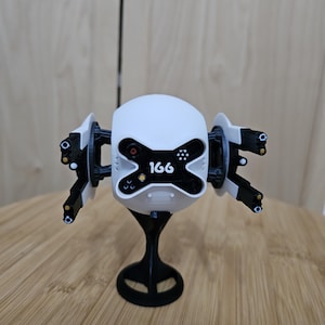May include: A white and black robotic figurine with the number "166" on the front. The figurine has multiple arms with circular details and is mounted on a black stand. The background is a light brown wooden surface.