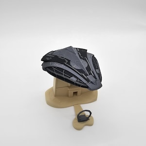 May include: A 3D printed model of a spaceship with a gray and black color scheme. The spaceship is sitting on a beige base with a small ring attached to the base.