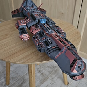 May include: A detailed model of a futuristic spaceship with a gray and copper color scheme. The spaceship has a long, slender body with a pointed nose and a series of fins and wings along its sides. The model is sitting on a wooden table.