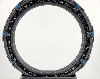 Può includere: Un modello stampato in 3D in nero e grigio di una Stargate, un dispositivo immaginario utilizzato per i viaggi interstellari. La Stargate è una struttura ad anello con un'apertura circolare al centro. L'anello è decorato con una serie di simboli e glifi. La Stargate è montata su una base nera.