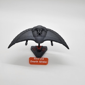 May include: A black plastic model of a Goa'uld Death Glider spaceship on a red stand. The stand has the words "Goa'uld Death Glider" printed on it.