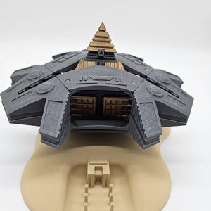 May include: A detailed 3D-printed model of a futuristic spacecraft or structure. The model features a dark gray body with intricate geometric designs and a central gold-colored pyramid-shaped structure. It rests on a tan-colored base with architectural elements.