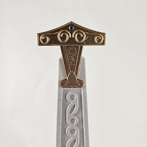 May include: A gold-colored decorative top with intricate designs and a blue jewel in the center, mounted on a white pillar with a repeating pattern.