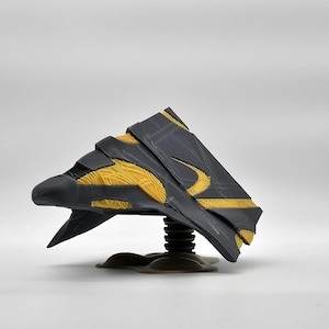 Puede incluir: Un juguete de nave espacial negro y amarillo con una base de resorte. La nave espacial tiene un diseño afilado y angular con detalles intrincados.