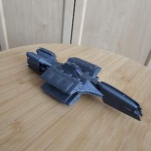 May include: A dark gray spaceship model with detailed features, including engines and wings. The model is made of plastic and is sitting on a wooden surface.