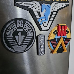 May include: A collection of five 3D printed Stargate SG-1 themed magnets. The magnets feature the Atlantis logo, SG-1 logo, SGC logo, a Goa'uld symbol, and a Jaffa symbol.