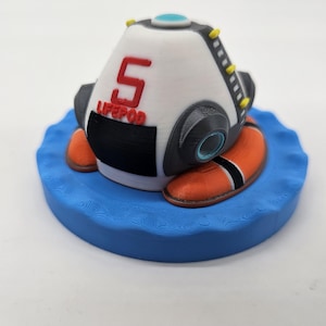 Könnte beinhalten: Ein 3D-gedrucktes Modell einer weißen und schwarzen Life Pod, mit der Zahl 5 und dem Wort "LIFEPOD" in Rot. Die Kapsel hat orangefarbene und schwarze Akzente und sitzt auf einer blauen, wellenförmigen Basis. Gelbe und blaue Details sind ebenfalls vorhanden.