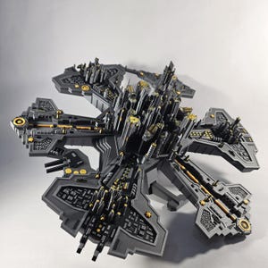 May include: A 3D printed model of a futuristic city, with a central tower and six arms extending outwards. The model is made of black plastic with gold accents.