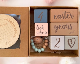 Może przedstawiać: Zestaw drewnianych klocków w brązowym pudełku. Klocki są pomalowane na różne kolory i mają napisy "easter", "years", "look who is", "4", "2" i symbol serca. Jest też drewniane kółko z napisem "I'm here" i miejscem na imię, datę, wagę i wzrost.