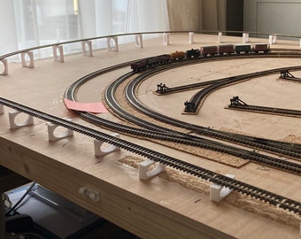 May include: A model train track layout with multiple tracks and a train on the outer track. The track is laid out on a wooden surface with white supports.