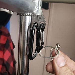 May include: A silver-colored metal bracket with the letter H engraved on it, attached to a vertical chrome pole. A black carabiner is clipped into the bracket, and a small loop of thin, light-colored cord is held near the carabiner attachment point.