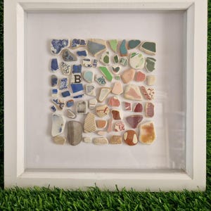 May include: A white framed artwork displaying a mosaic of ceramic fragments. The fragments are in various shapes and sizes, with blue, green, red, and white colours. The frame is white, and the artwork is set against a white background.